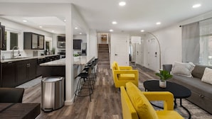 Eine moderne Küche mit dunklen Schränken und einer zentralen Kochinsel, ein Essbereich mit gelben Stühlen und ein Wohnzimmer mit einer Couch und einem Couchtisch.