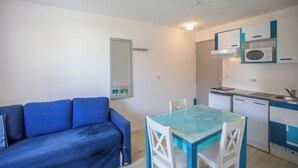 Un espace de vie compact comprenant un canapé bleu, une table à manger avec un plateau en verre, des chaises blanches, un four à micro-ondes, un petit évier et une kitchenette compacte.