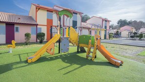 Un terrain de jeux avec des toboggans et des structures d’escalade, situé devant une rangée de maisons colorées aux toits recouverts de tuiles rouges.