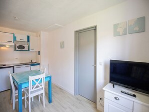 A compact kitchen with a microwave, sink, and a dining area with a table and chairs. A closed door is visible, and a TV is mounted on a cabinet.