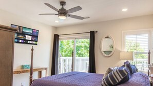 A bedroom with a bed, a ceiling fan, a TV mounted on the wall, a window with curtains, and a view of greenery outside.