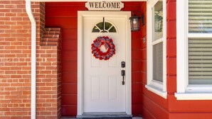 A red brick building with a white door, a 'WELCOME' sign above the door, and a wreath on the door.