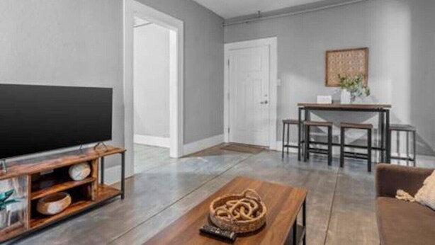 Een moderne woonkamer met een flatscreen televisie op een houten standaard, een salontafel met een geweven decoratie, een bruine bank en een eethoek met een kleine tafel en stoelen.
