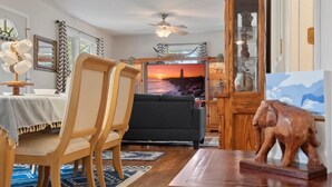 A living room with a sofa, wooden chairs, a dining table with a chandelier, a television displaying a sunset, and a wooden cabinet with a decorative elephant. - Modern Bungalow Home with Mid-Century Flair (Springfield)