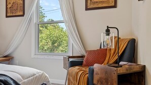 A bedroom with a bed, a wooden side table, a chair with a cushion, a window with curtains, and framed artwork on the wall. - Modern Bungalow Home with Mid-Century Flair (Springfield)