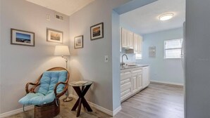 A room with a wicker chair, a small table, and a lamp. There is a kitchen area with white cabinets and a window.