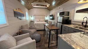 A compact, modern living space with a kitchenette, a sofa, a bed, and a television. - Tiny Home Retreat Near Deep Creek • Hot Tub •Fenced Yard •Perfect Couples Escape (Bryson City)