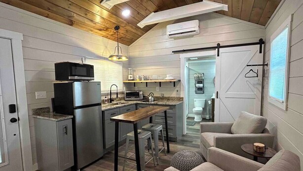 A compact kitchen with modern appliances, a dining area with stools, and a cozy seating area with a sofa and a small table. - Tiny Home Retreat Near Deep Creek • Hot Tub •Fenced Yard •Perfect Couples Escape (Bryson City)