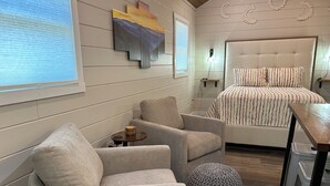 A bedroom with a bed, two armchairs, a small round ottoman, a bedside table, and a wall-mounted artwork. - Tiny Home Retreat Near Deep Creek • Hot Tub •Fenced Yard •Perfect Couples Escape (Bryson City)