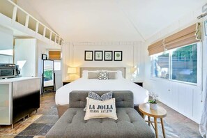 A hotel room with a bed, a desk, a chair, a small table, and a window with blinds. - Secluded Windansea Beach Rental Cottage (La Jolla)