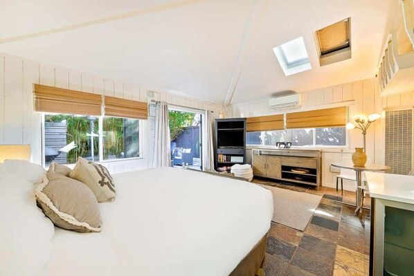 A bedroom with a large bed, a TV, a kitchenette, and a sliding glass door leading to an outdoor area. - Secluded Windansea Beach Rental Cottage (La Jolla)