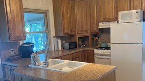 A kitchen with wooden cabinets, a white refrigerator, a microwave, a stove, and a sink. There are two wicker bar stools and a ceiling light fixture. - Beautiful cottage on the lake
2 bedroom, 1 bath, sleeps 7-8 (Three Rivers)
