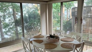 A dining area with a round table, white chairs, and large windows offering a view of trees and a deck. - Beautiful cottage on the lake
2 bedroom, 1 bath, sleeps 7-8 (Three Rivers)