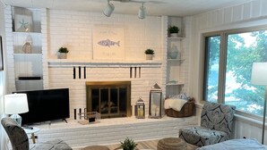 A modern living room with a fireplace, white brick walls, and a large window. - Beautiful cottage on the lake
2 bedroom, 1 bath, sleeps 7-8 (Three Rivers)
