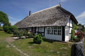 A traditional thatched-roof cottage with white walls and black trim, surrounded by a garden with flowering plants and a stone pathway. - Idyllic seaside location close to the beach, private bathing area - Gutshof Ostseeblick FeWo 04 - (Putbus)