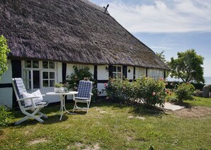 Una cabana amb teulada de palla i façana blanca, amb dues cadires blanques, una taula petita i un jardí ple de flors roses.