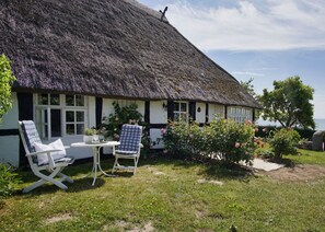 A thatched-roof cottage with a white exterior, two white chairs, a small table, and a garden with pink flowers.