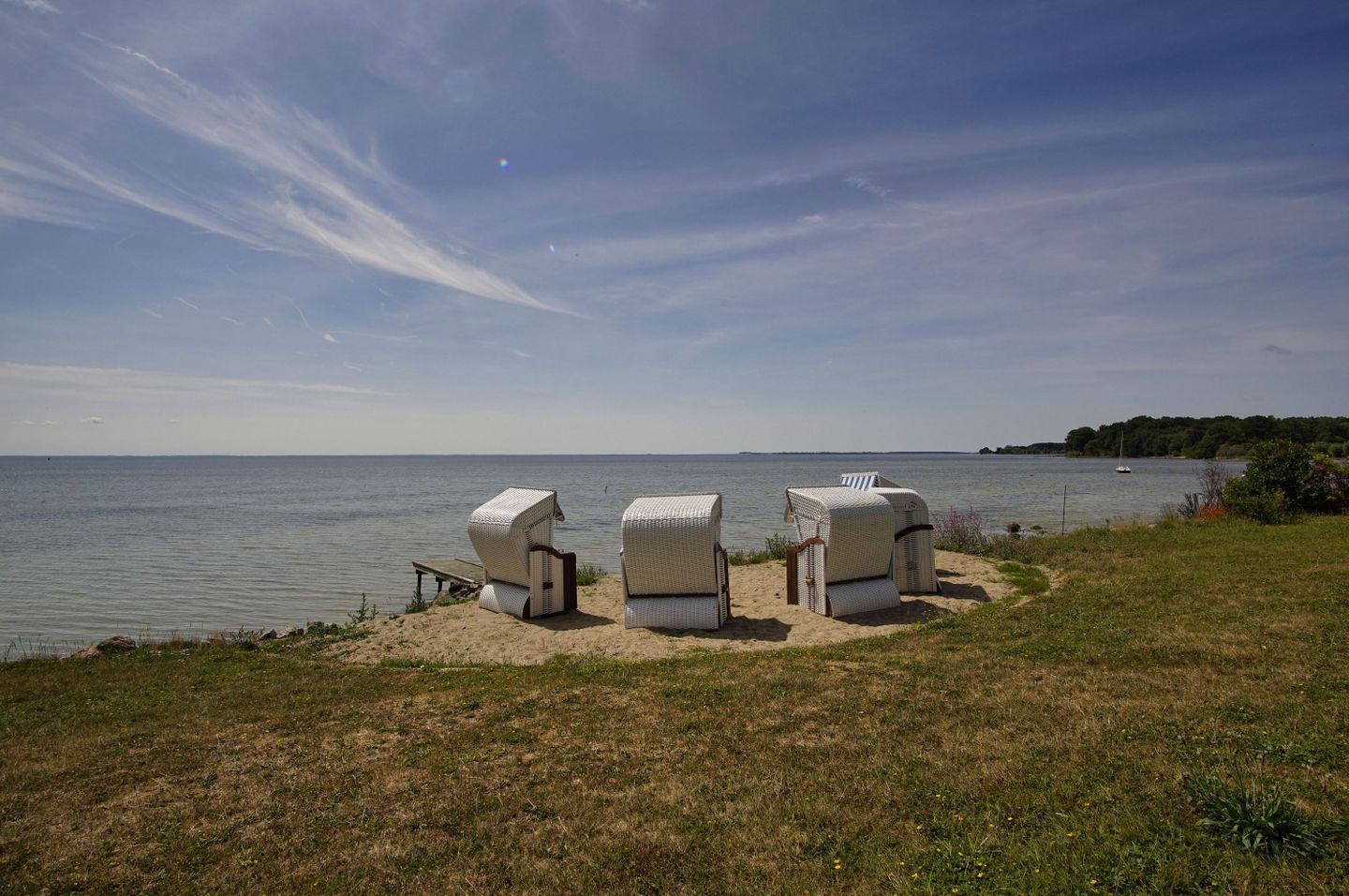 Een strand met vier witte rieten stoelen en een tafeltje, met uitzicht op een rustige zee en een verre kustlijn.