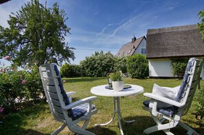Two rocking chairs with checkered cushions, a round table with a potted plant, and a house with a thatched roof in the background. - Idyllic seaside location close to the beach, private bathing area - Gutshof Ostseeblick FeWo 04 - (Putbus)