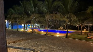 Vista nocturna de una zona de piscina con palmeras y una estructura iluminada en azul al fondo.