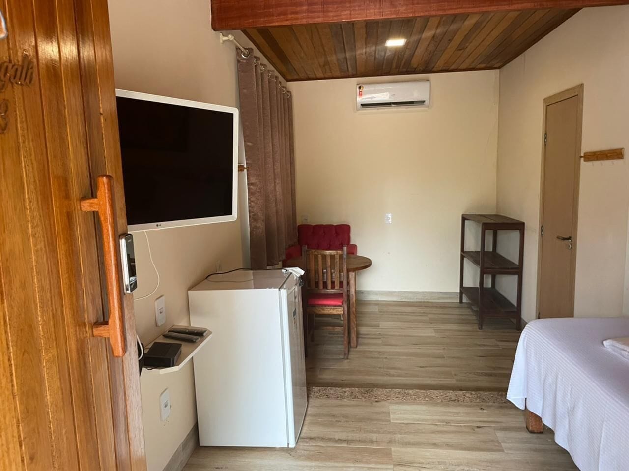 Habitación compacta con techo de madera, refrigerador blanco, televisor montado en la pared, pequeña mesa de madera con una silla y cama con cubrecama blanco.