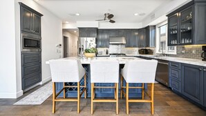 A modern kitchen with dark cabinetry, a central island with bar stools, a built-in oven, and a ceiling fan.