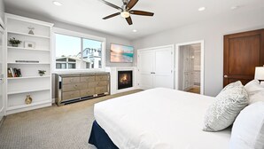 A bedroom with a large bed, a fireplace, a ceiling fan, and a view of a house through a window.