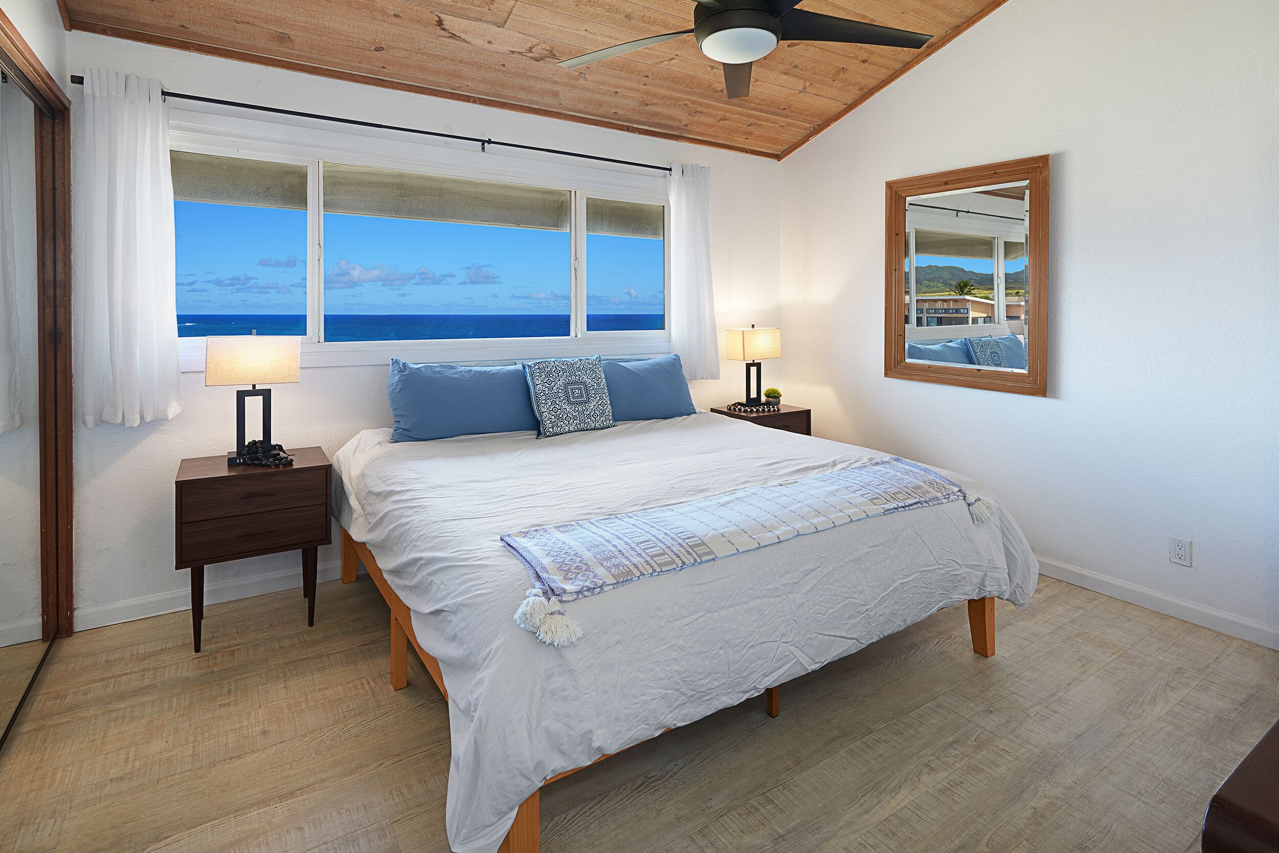 Un dormitorio con una cama grande, dos mesitas de noche, un ventilador de techo, una ventana con vistas al mar y un espejo en la pared.