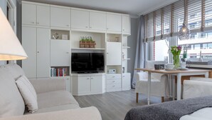 Ein modernes Wohnzimmer mit Flachbildfernseher, weißer Möbel und einem Essbereich mit Tisch und Stühlen.