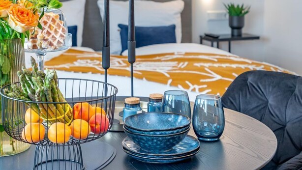 A modern hotel room with a round dining table, black chairs, a blue pendant light, and a bed with a brown and white patterned bedspread. - Schickes Stranddomizil in der 3. Etage, in Direkter Lage zum Strand - Andreas-dirks-straße 5 (Sylt)
