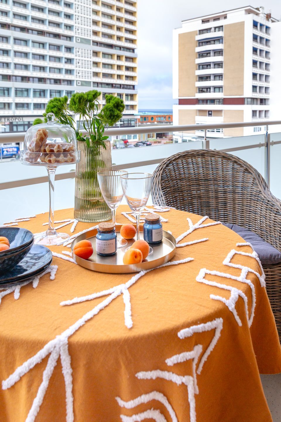 A table set with a tray of food, a vase with greenery, and a bottle of perfume on a balcony with a view of buildings.