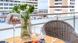 A table set with a tray of food, a vase with greenery, and a bottle of perfume on a balcony with a view of buildings. - Schickes Stranddomizil in der 3. Etage, in Direkter Lage zum Strand - Andreas-dirks-straße 5 (Sylt)