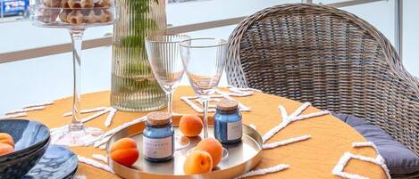 A table set with a tray of food, a vase with greenery, and a bottle of perfume on a balcony with a view of buildings.