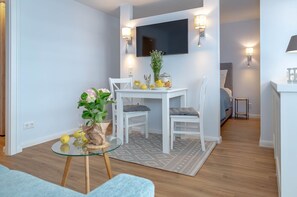 A modern living room with a glass coffee table, white dining table, and chairs. A flat-screen TV is mounted on the wall above the dining area.