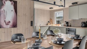 A modern dining area with a wooden table set for six, surrounded by black chairs. A large painting of a horse is on the wall, and the room has a minimalist design with a visible kitchen area in the background. - Bjørkstue by Interhome (Eikerapen)