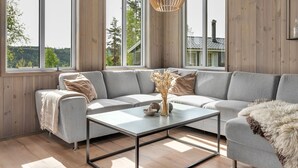 A modern living room with a grey sectional sofa, a glass coffee table, and large windows offering a view of trees and a building. - Bjørkstue by Interhome (Eikerapen)