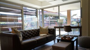 A modern living room with a leather sofa, a coffee table, and a view of a building through large windows. - Optimal Camelback, Scottsdale Luxury Condo Living All Year Round! (Scottsdale)
