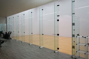 A racquetball court with glass walls and a wooden floor, surrounded by exercise equipment.