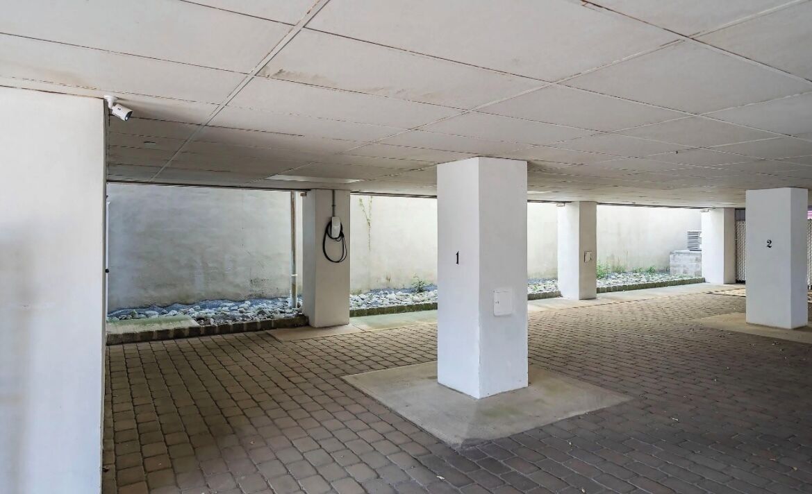 An indoor parking area with concrete pillars, a tiled floor, and a view of a landscaped area outside.