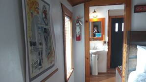 A room with a wooden ceiling, a bed with a floral bedspread, a small kitchenette, and a bathroom with a toilet and sink.