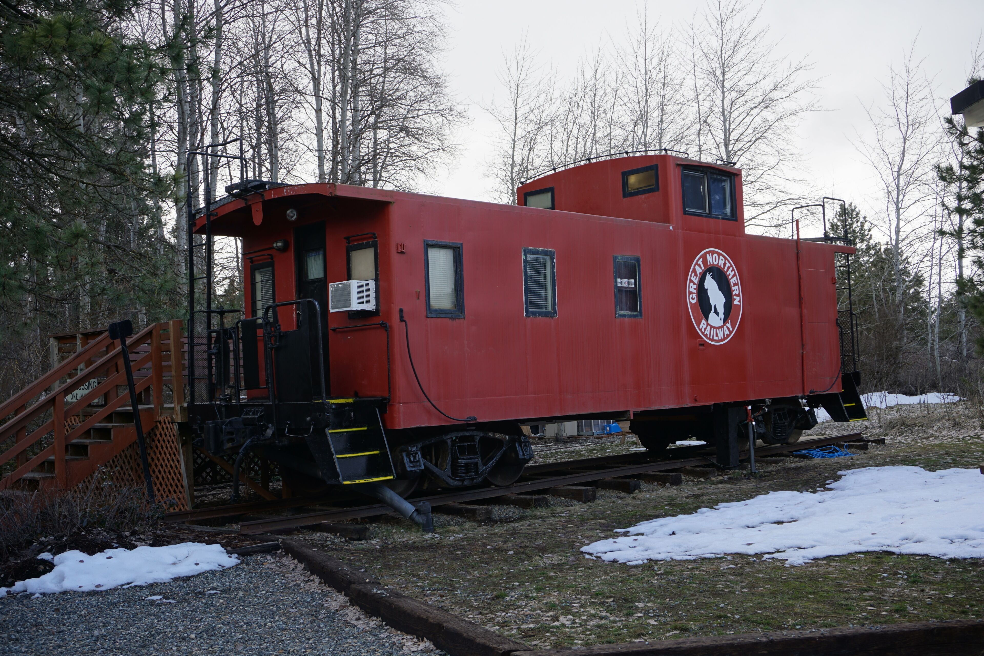 A red train caboose with a Great Northern Railway logo, situated on tracks with a wooden staircase leading up to it, surrounded by trees and snow.