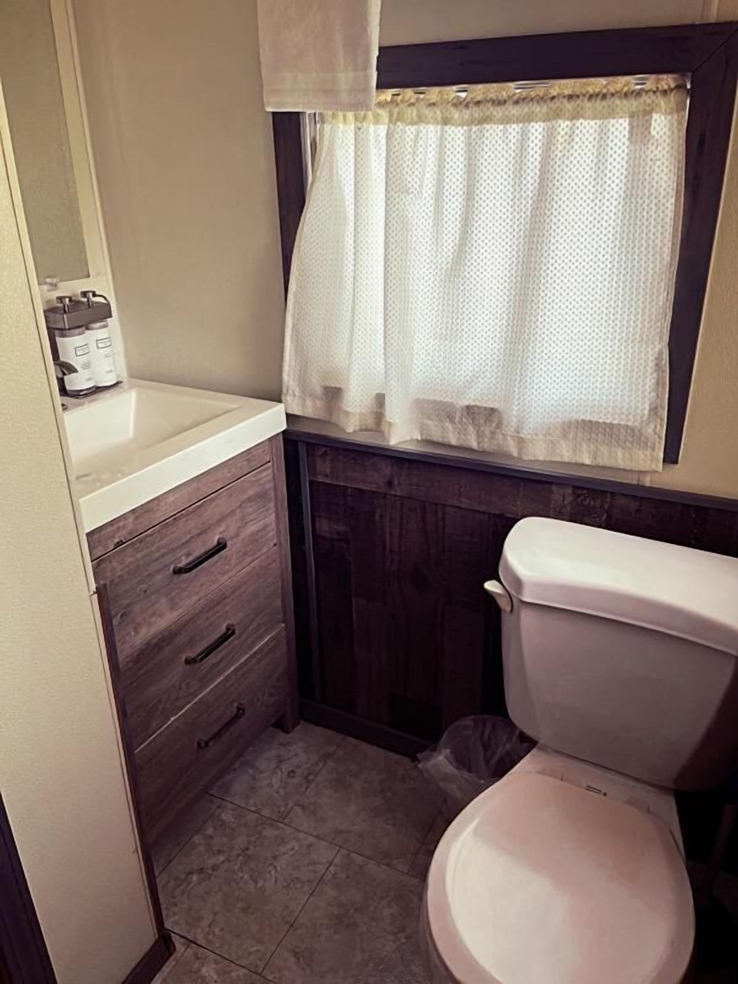 A small bathroom with a toilet, a wooden cabinet with drawers, and a window with a white curtain.
