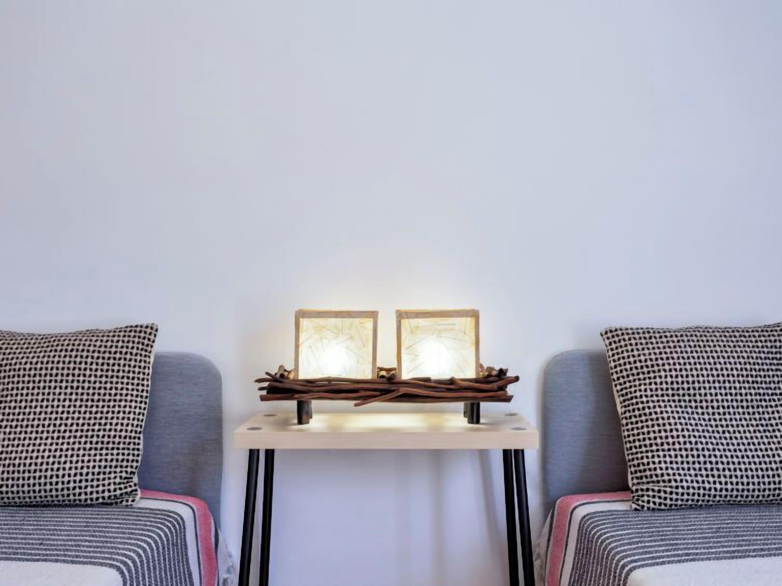 A room with two grey sofas, a small white table with two framed pictures, and a simple wall-mounted light fixture.
