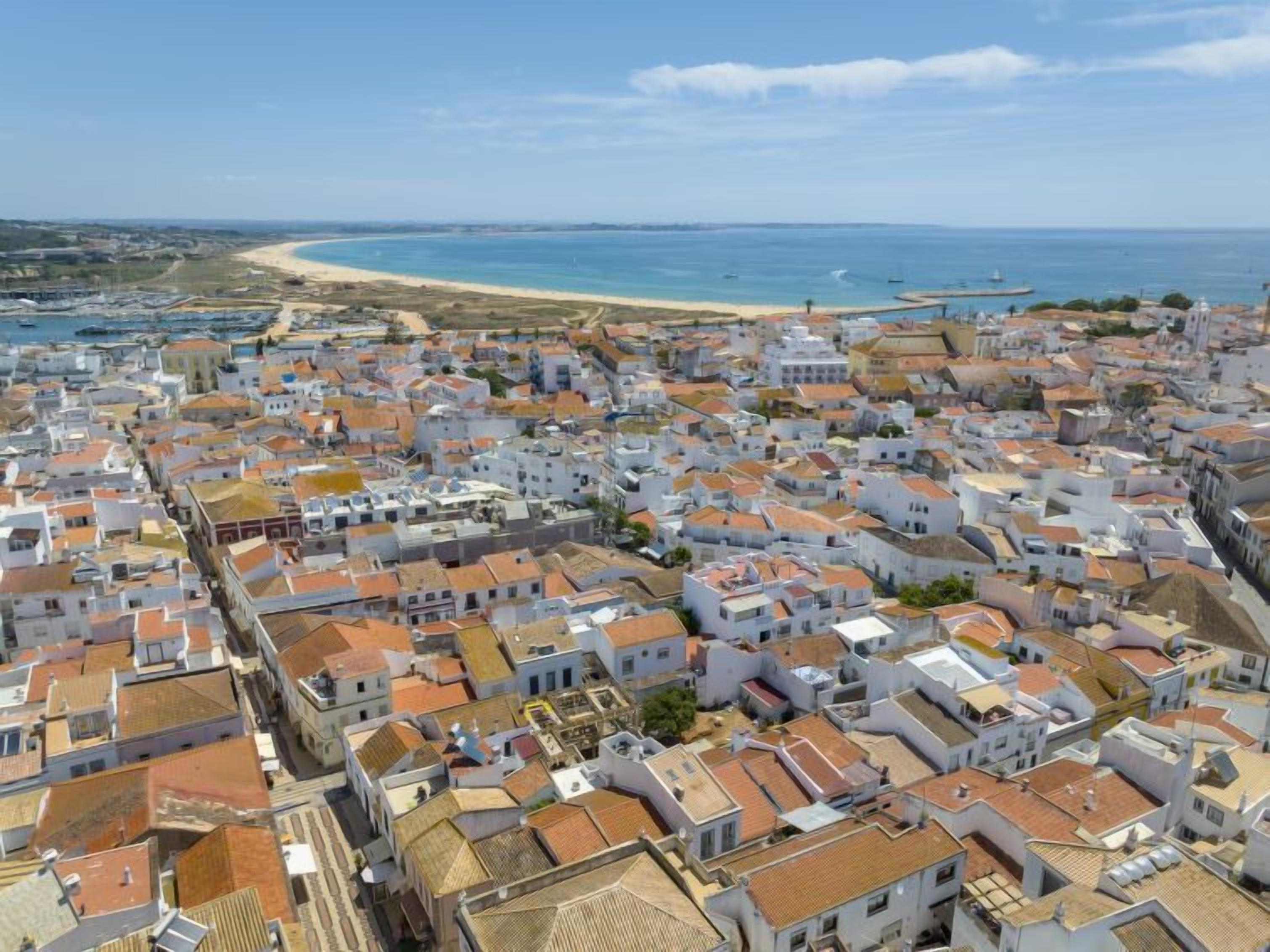 An aerial view of a coastal town with white buildings and red roofs, a beach, and a lighthouse in the distance.