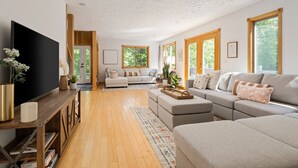 Un salon spacieux doté d’un grand canapé d’angle, d’un téléviseur à écran plat, de parquet et de grandes fenêtres laissant entrer la lumière naturelle.
