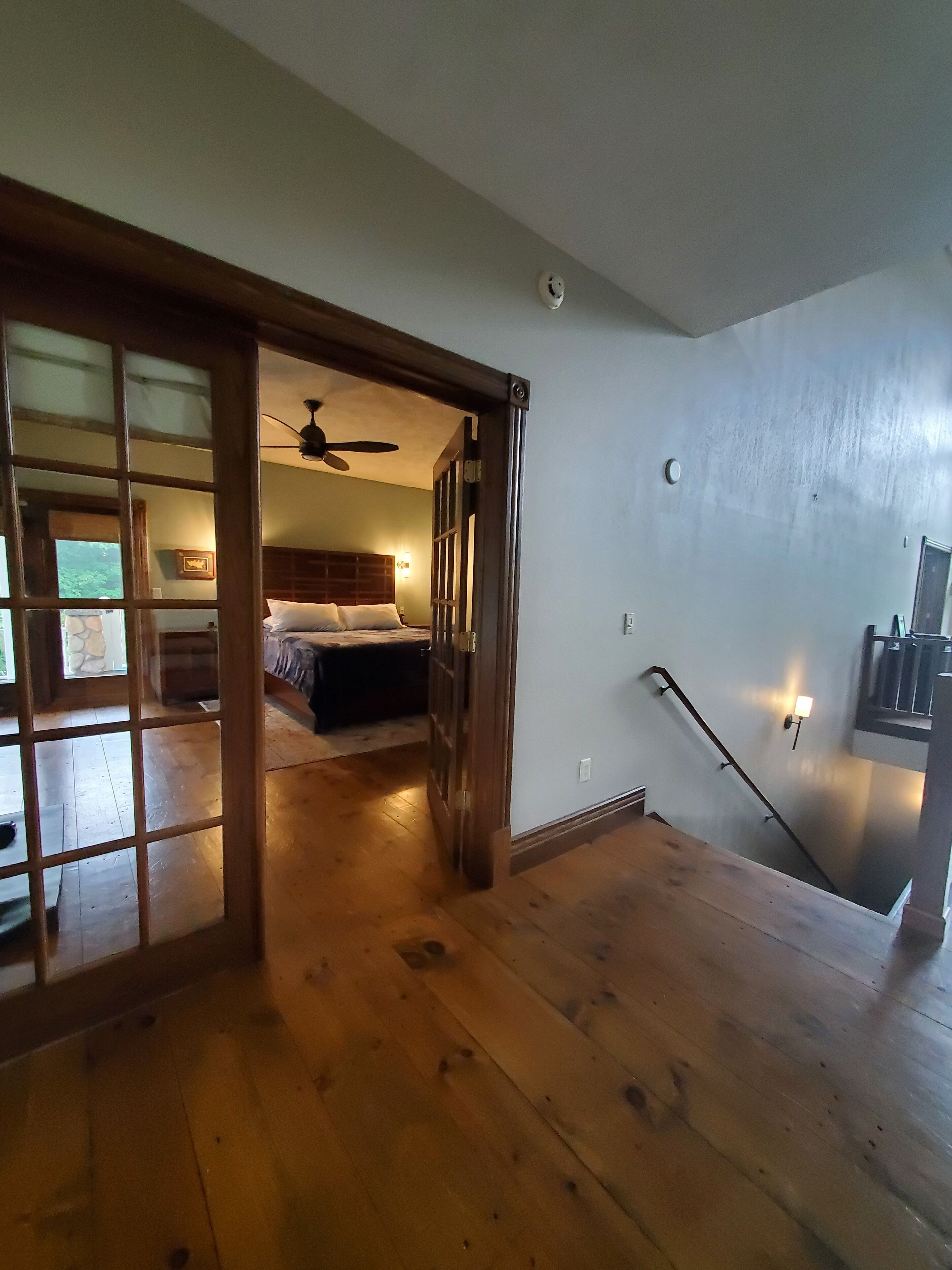 Une pièce spacieuse avec un plancher en bois, un lit doté d’une tête de lit, un ventilateur de plafond et un escalier muni d’une main courante.