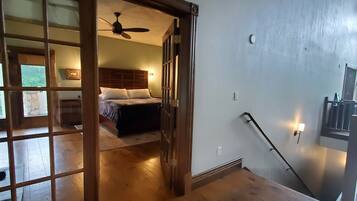 Une pièce spacieuse avec un plancher en bois, un lit doté d’une tête de lit, un ventilateur de plafond et un escalier muni d’une main courante.