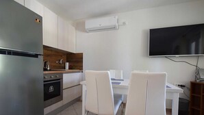 Une cuisine compacte équipée d’un réfrigérateur, d’une cuisinière et d’un four à micro-ondes. On y trouve une table à manger avec des chaises blanches et un téléviseur à écran plat fixé au mur.