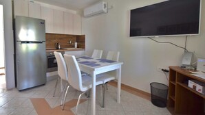 Une cuisine compacte équipée d’un réfrigérateur, d’une cuisinière et d’un four à micro-ondes. On y trouve une table à manger avec des chaises, ainsi qu’un grand téléviseur à écran plat fixé au mur.
