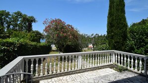 Un balcon dallé de pierres, doté d’une balustrade blanche, entouré d’une végétation luxuriante et d’un grand arbre à feuilles persistantes.